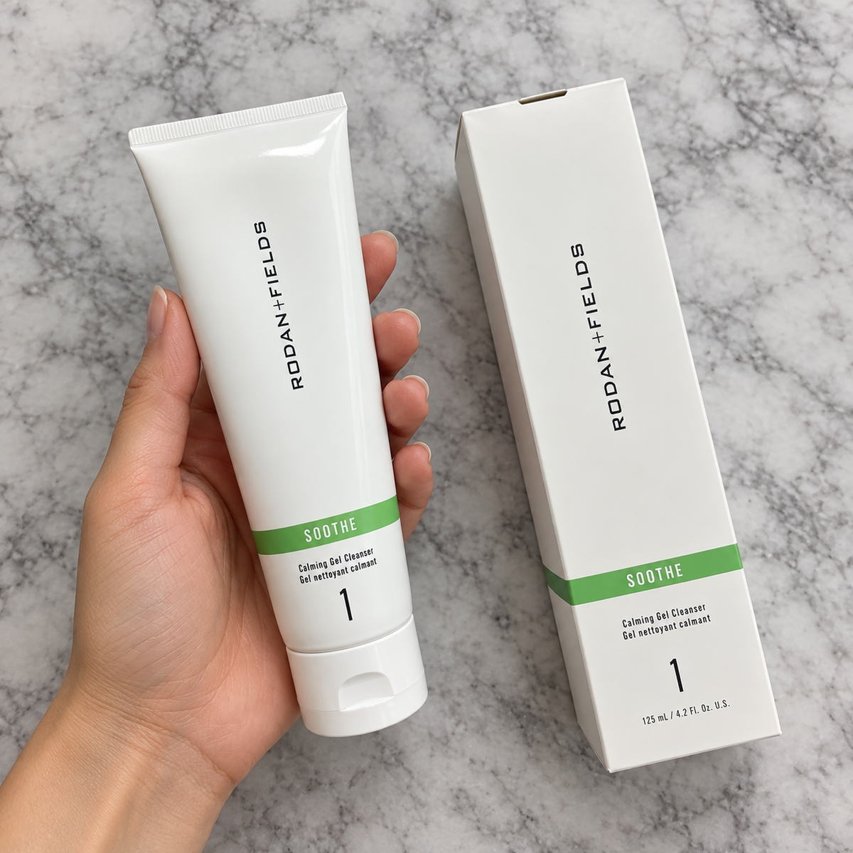 Overhead view of a hand holding the complete SOOTHE cleanser tube, showing its green label band and closed flip-cap beside the box.