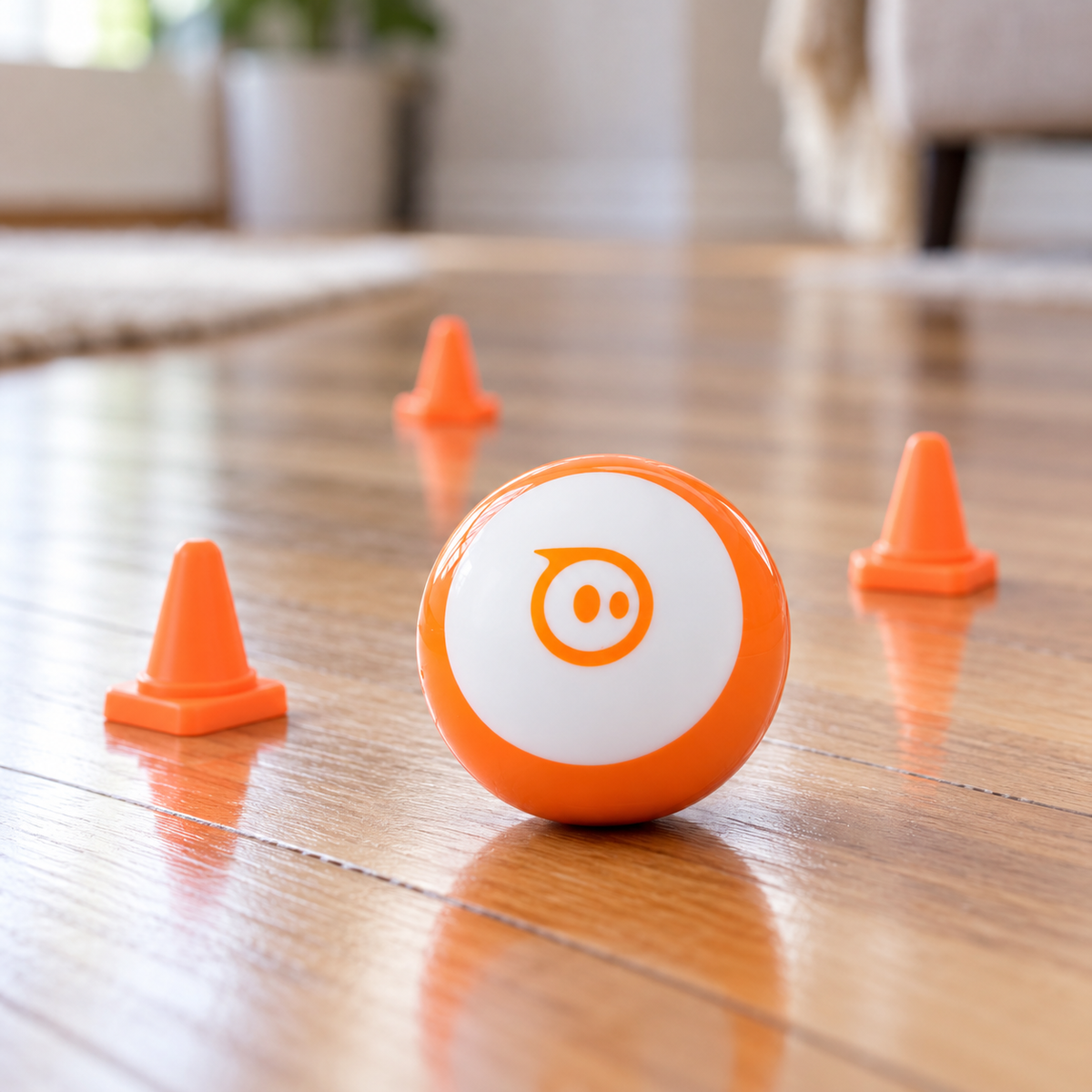 Orange and white Sphero Mini robotic ball beside three traffic cones on a hard indoor floor.