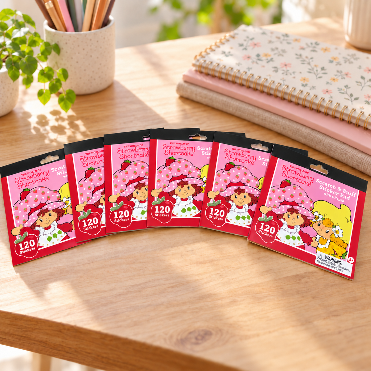 AI-generated lifestyle scene showing six closed Strawberry Shortcake scratch-and-sniff sticker pads,120 stickers per pad, on a craft desk. Props not included.