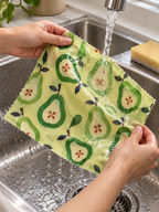 AI-generated lifestyle illustration: Green pear-print SuperBee wrap being gently rinsed; food and props not included.