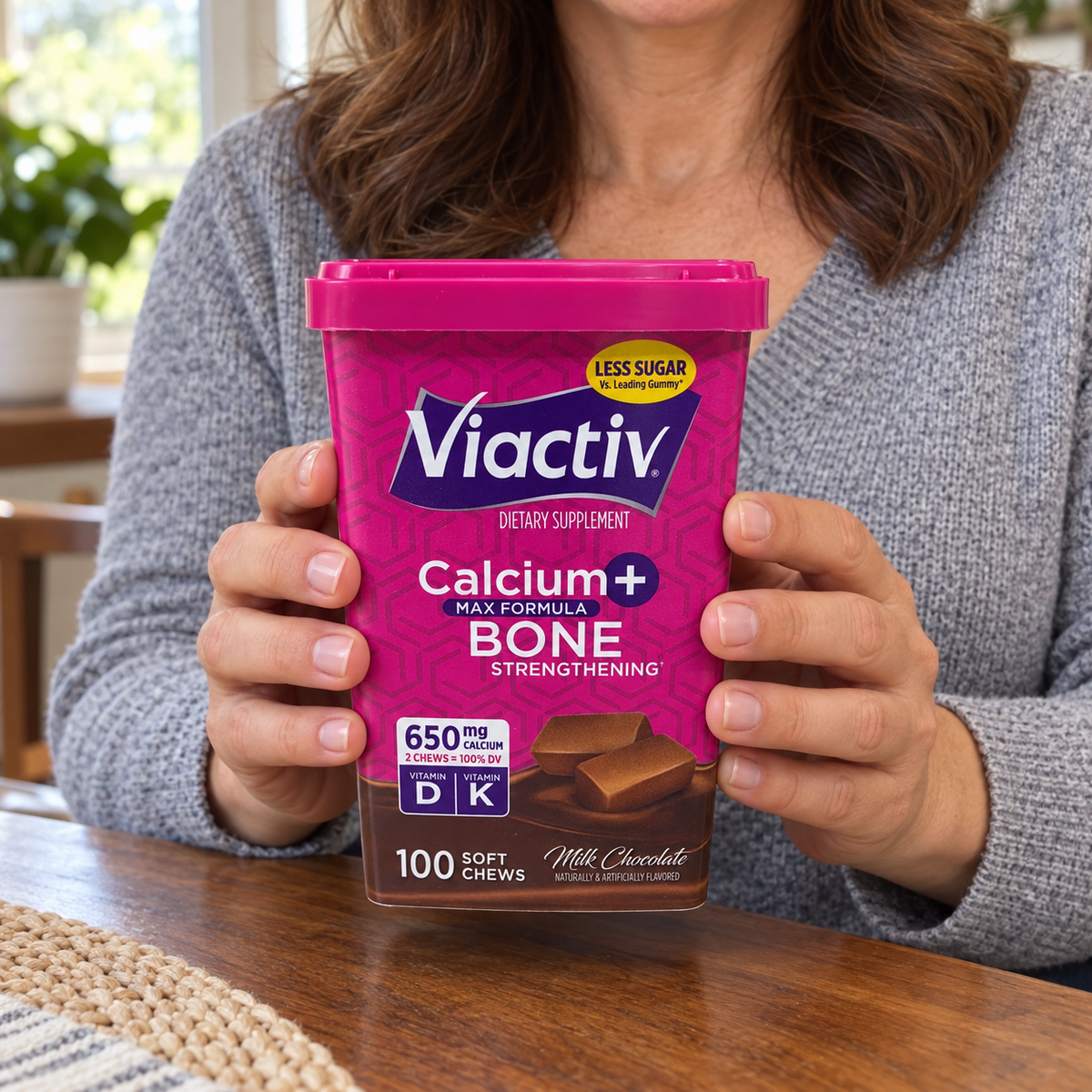An adult holding a closed Viactiv Milk Chocolate 100-chew container at a table.