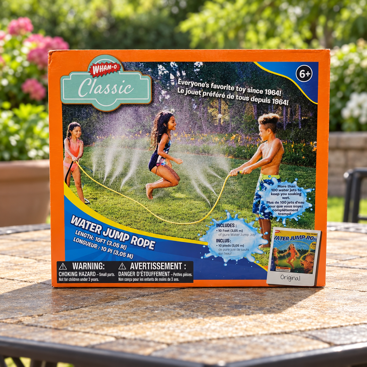 AI-generated lifestyle scene of one closed Wham-O Classic Water Jump Rope box on a patio table. People and water appear only in printed packaging artwork; props not included.